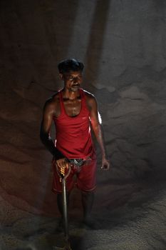 A man in a red tank top stands with a shovel under dramatic lighting, suggesting labor.
