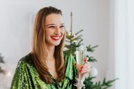在室内圣诞树旁，一位身穿绿色连衣裙的妇女手持香槟杯，面带微笑