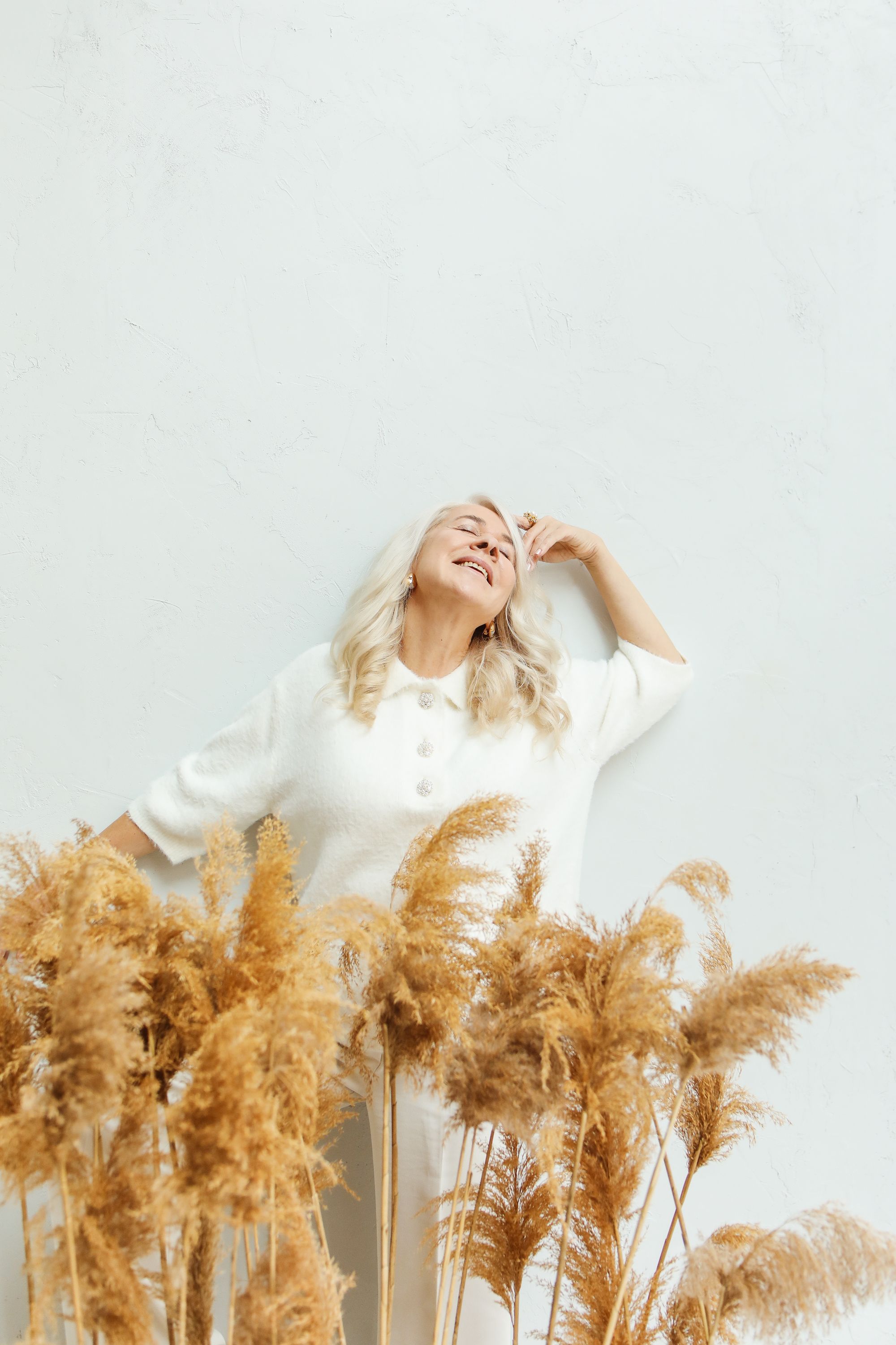 Senior woman in white smiling amidst golden pampas grass in a minimalist setting.