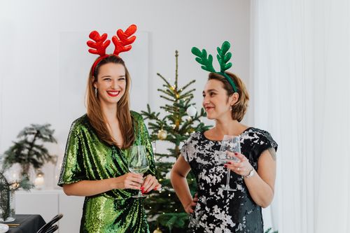Free Two women wearing reindeer headbands celebrate with wine by a Christmas tree. Stock Photo
