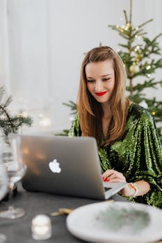 Smiling woman in a green dress using a laptop at a Christmas-decorated table.