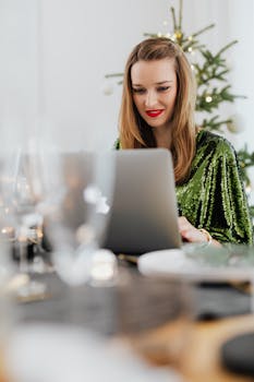 一位身穿绿色连衣裙的女子面带微笑，在室内圣诞树旁使用笔记本电脑，气氛喜庆
