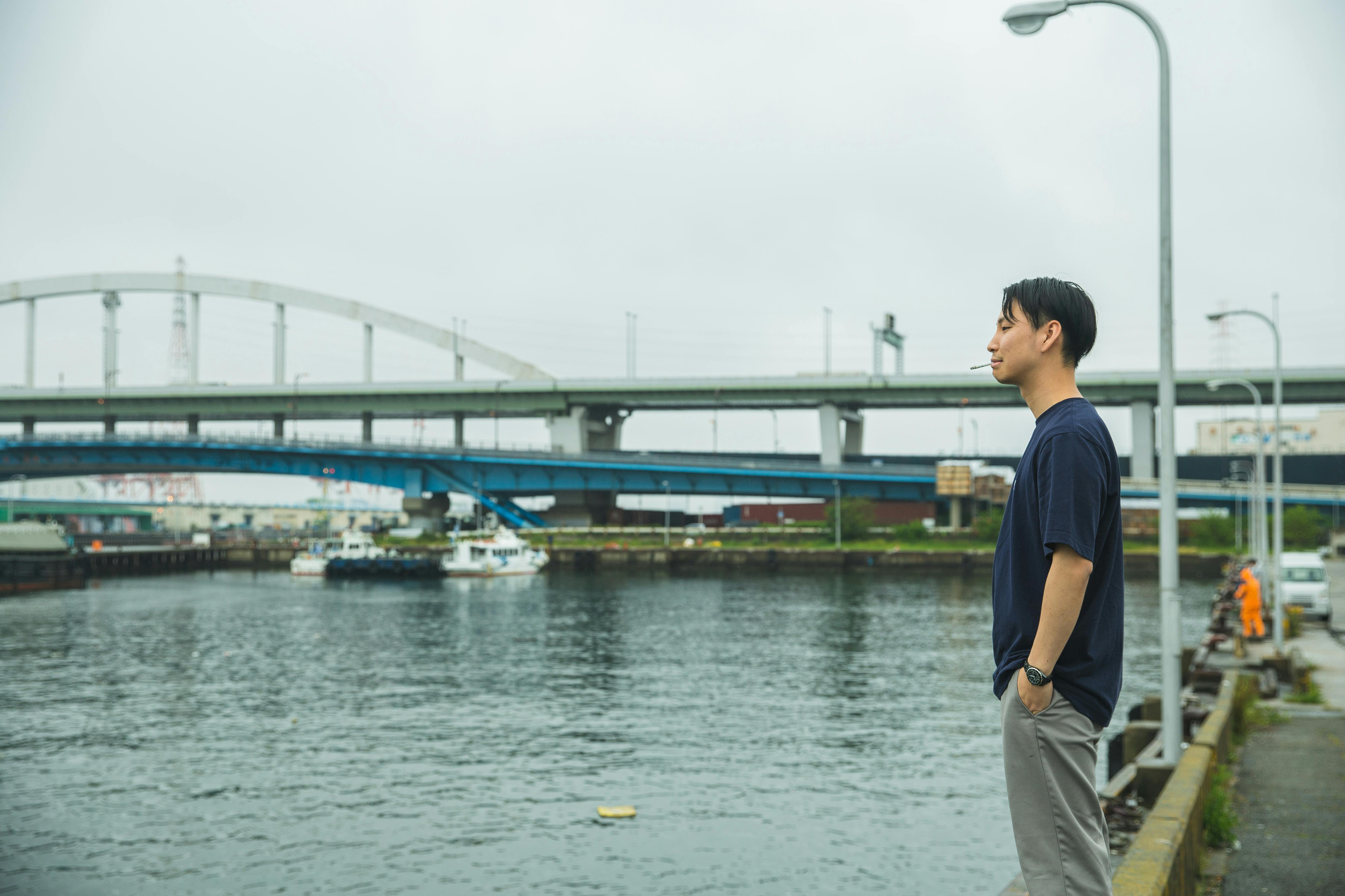 堤岸上，一位穿着休闲装的亚洲年轻人手插口袋，侧身而立，陷入沉思
