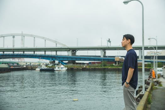堤岸上，一位穿着休闲装的亚洲年轻人手插口袋，侧身而立，陷入沉思