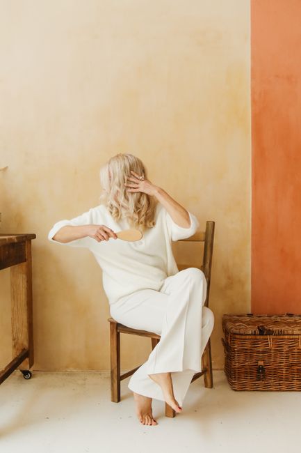 Elegant senior woman with white hair in cozy white attire, sitting indoors, brushing hair.