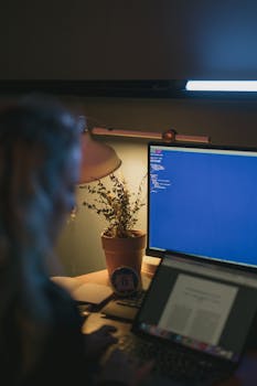 A person using a computer and laptop at night with a potted plant and desk lamp.