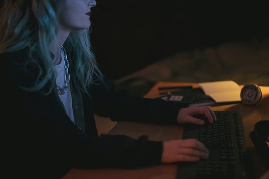 Woman typing on a keyboard at night, illuminated by screen light.