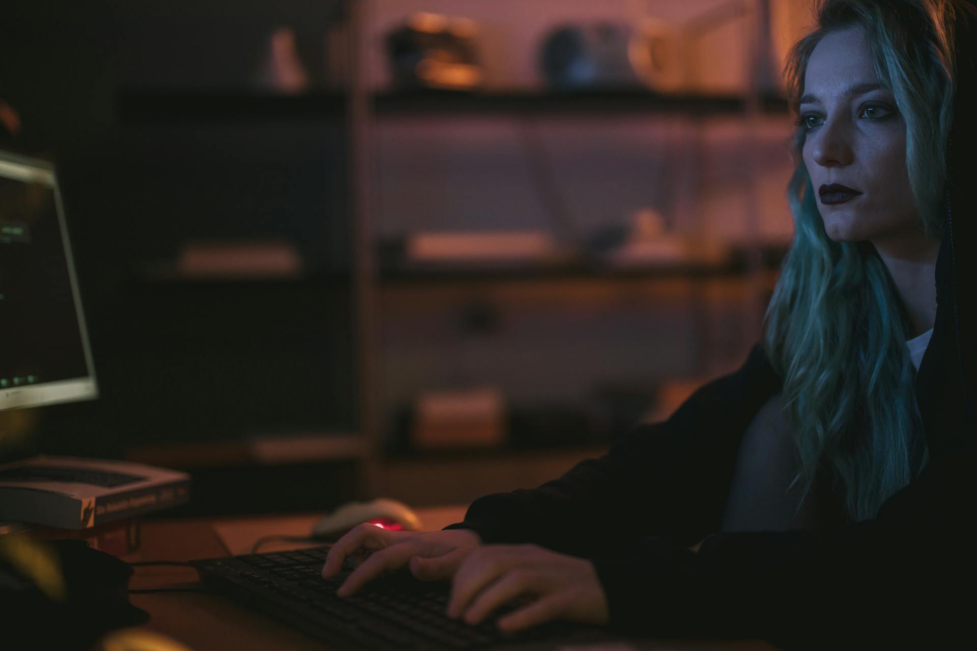 A woman with blue hair types on a keyboard in a dark, tech-themed room, implying cybersecurity work.