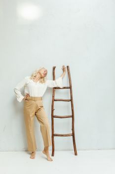 一位时尚的高级女性赤脚倚靠在极简主义的白墙木梯旁