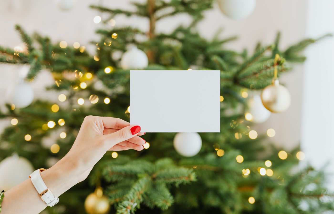 Hand holding a blank card with a decorated Christmas tree in the background, perfect for holiday greetings.