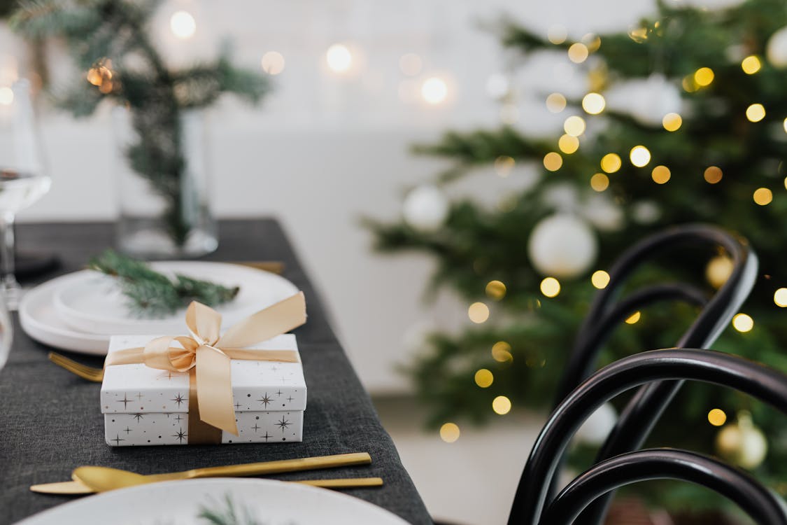 White gift box with a gold ribbon on a festive dinner table next to a decorated Christmas tree.