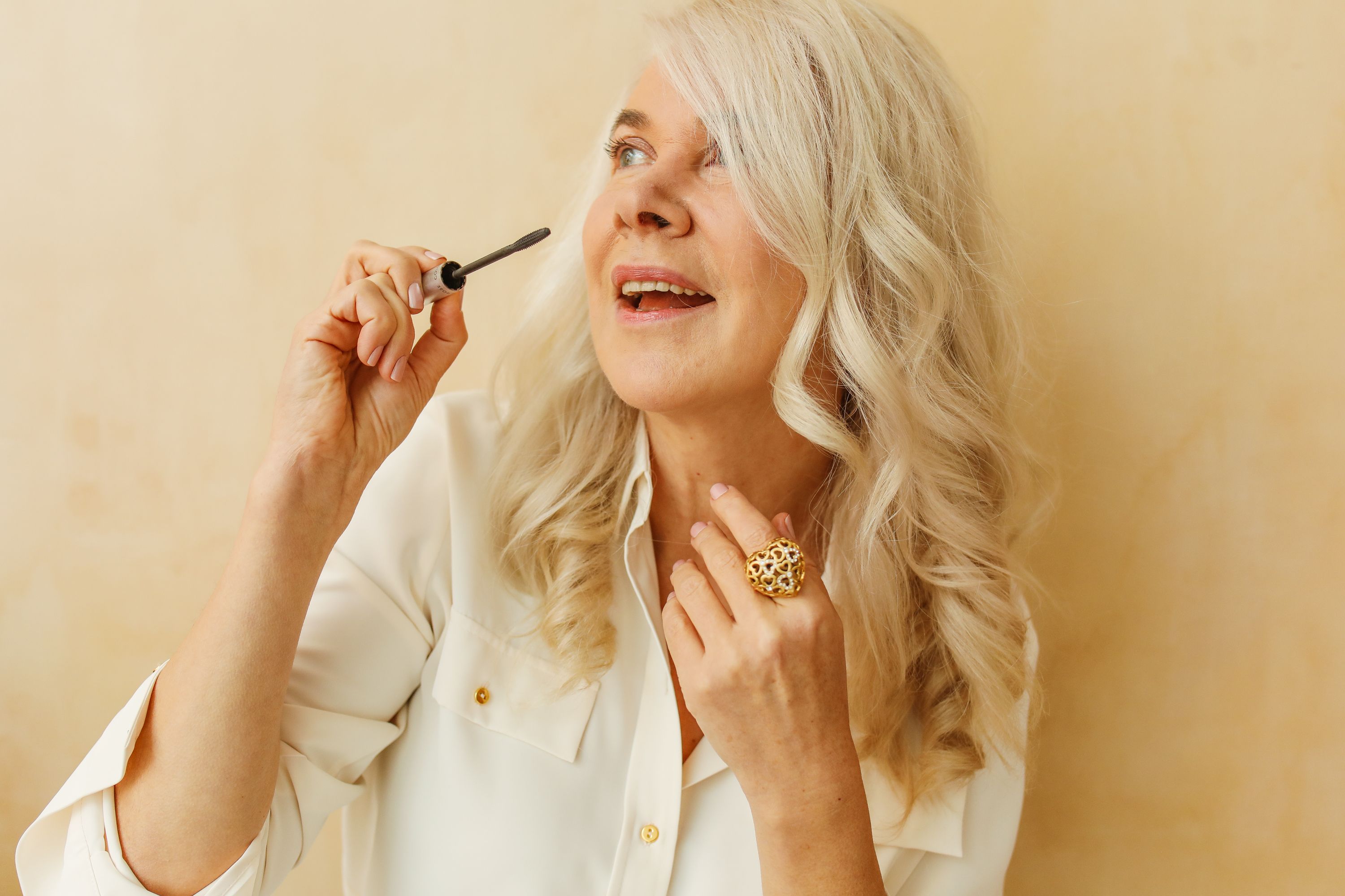 Joyful senior woman applying mascara with a smile in a bright, stylish setting.