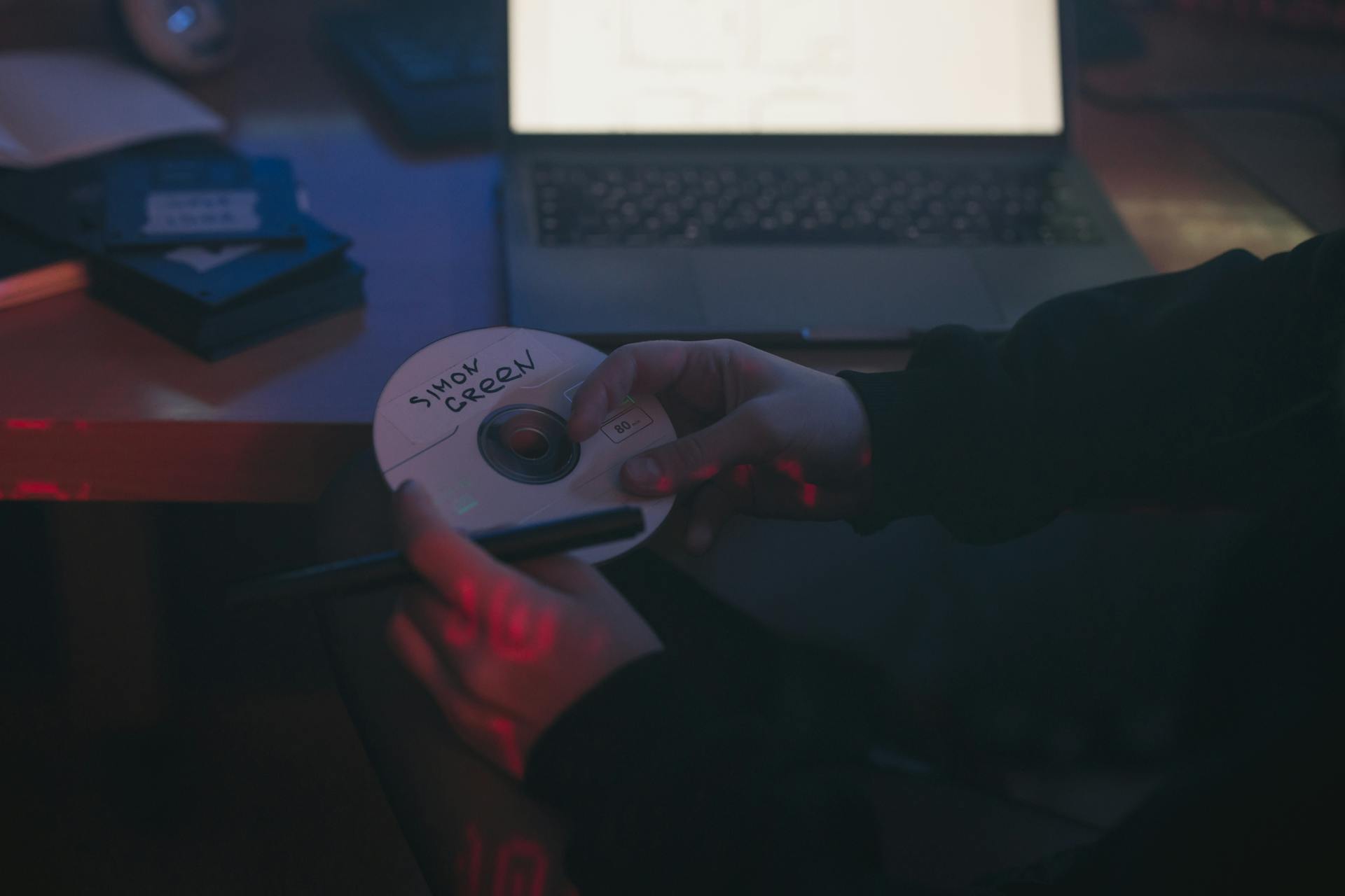 A person holds a CD labeled 'Snow Green' in a dimly lit room with a laptop.