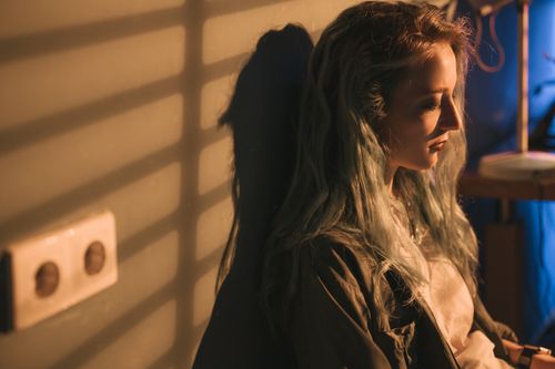 Free A woman with long hair leans against a wall, illuminated by sunlight streaming through blinds. Stock Photo