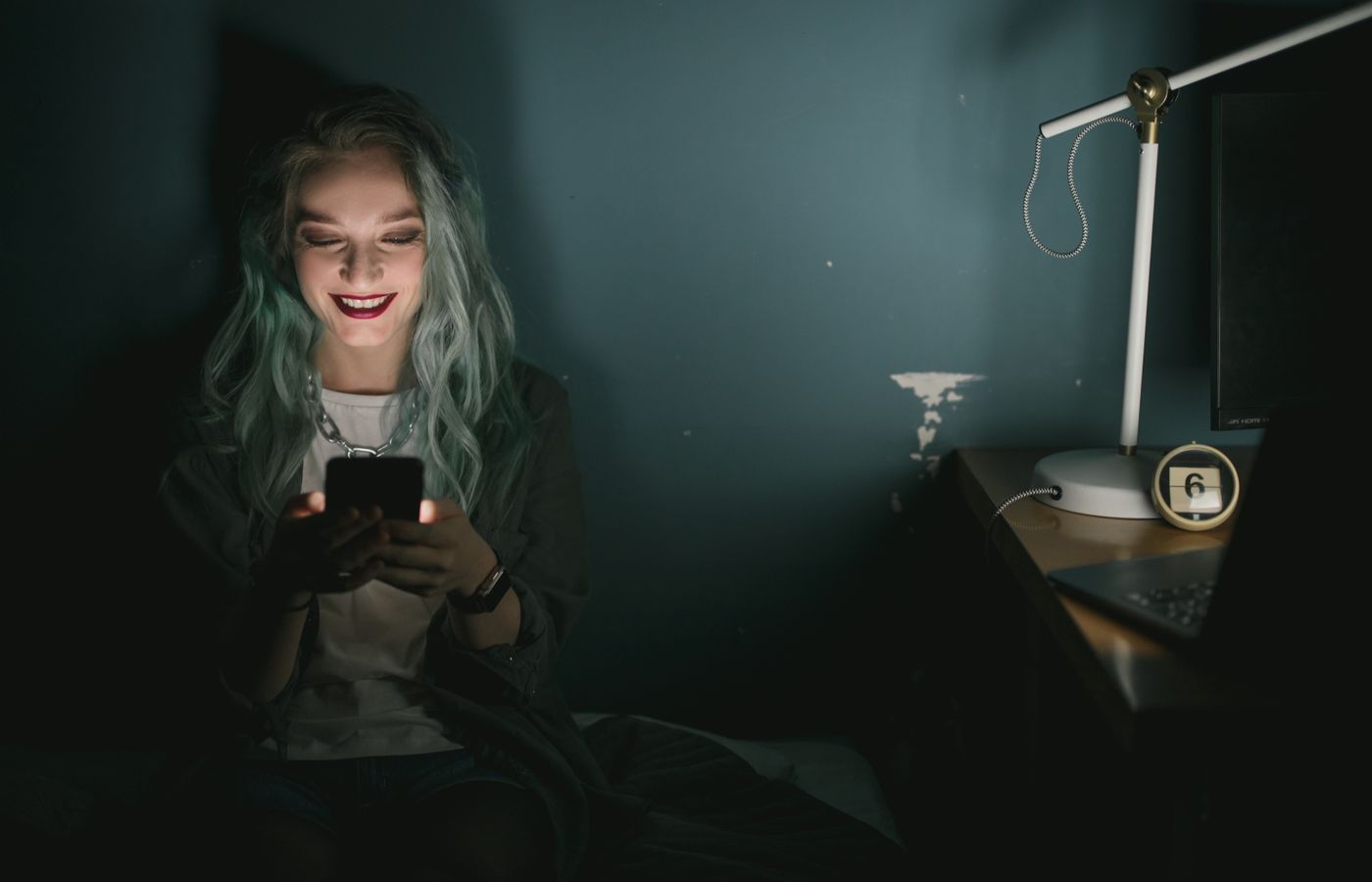 A young woman smiles at her smartphone in a dimly lit room, illuminated by the screen light.