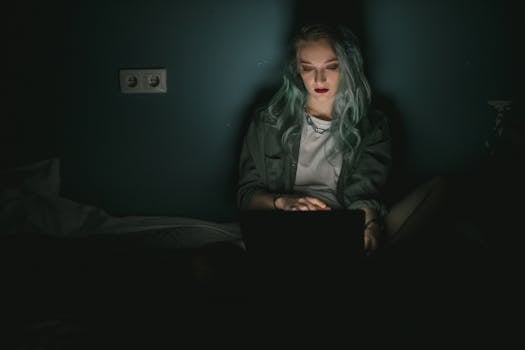 A woman with dyed hair using a laptop in a dimly lit room, working late at night.