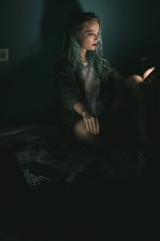A young woman with dyed hair uses her phone in a dark room, illuminated by the screen light.