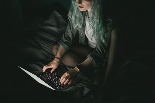一名染着彩色头发的年轻女子在黑暗中操作笔记本电脑，屏幕投射出柔和的光芒