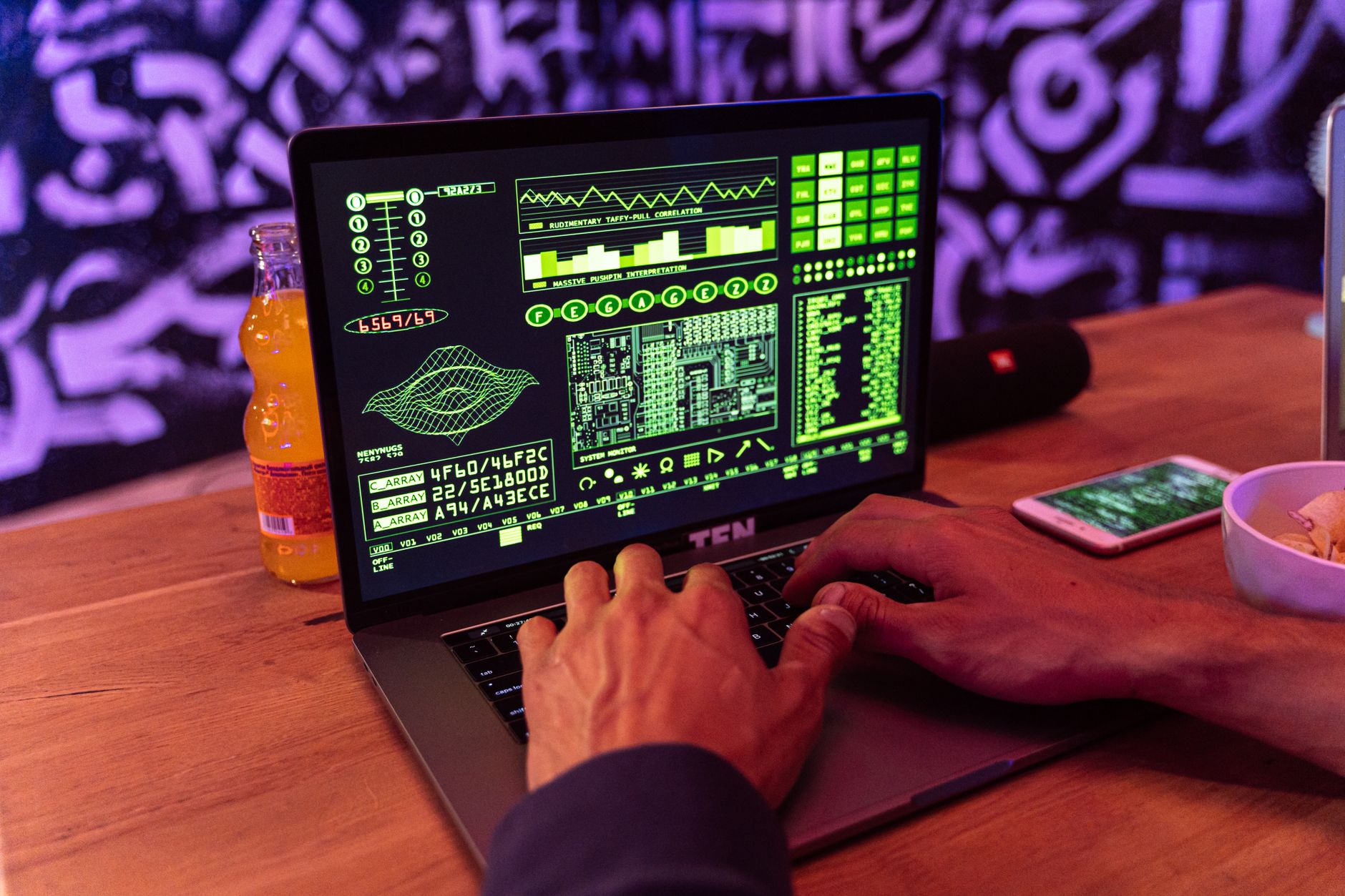 A person typing on a laptop displaying green code with an orange bottle and smartphone nearby.