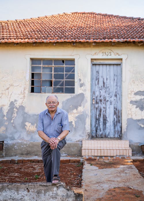 Senior male with gray hair in eyeglasses standing near aged building with weathered walls and looking at camera