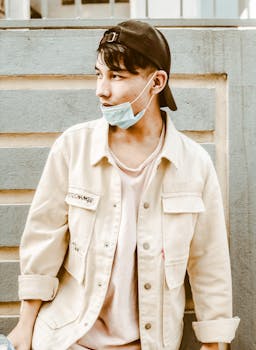 A young man wearing a face mask and casual attire poses against a textured wall.