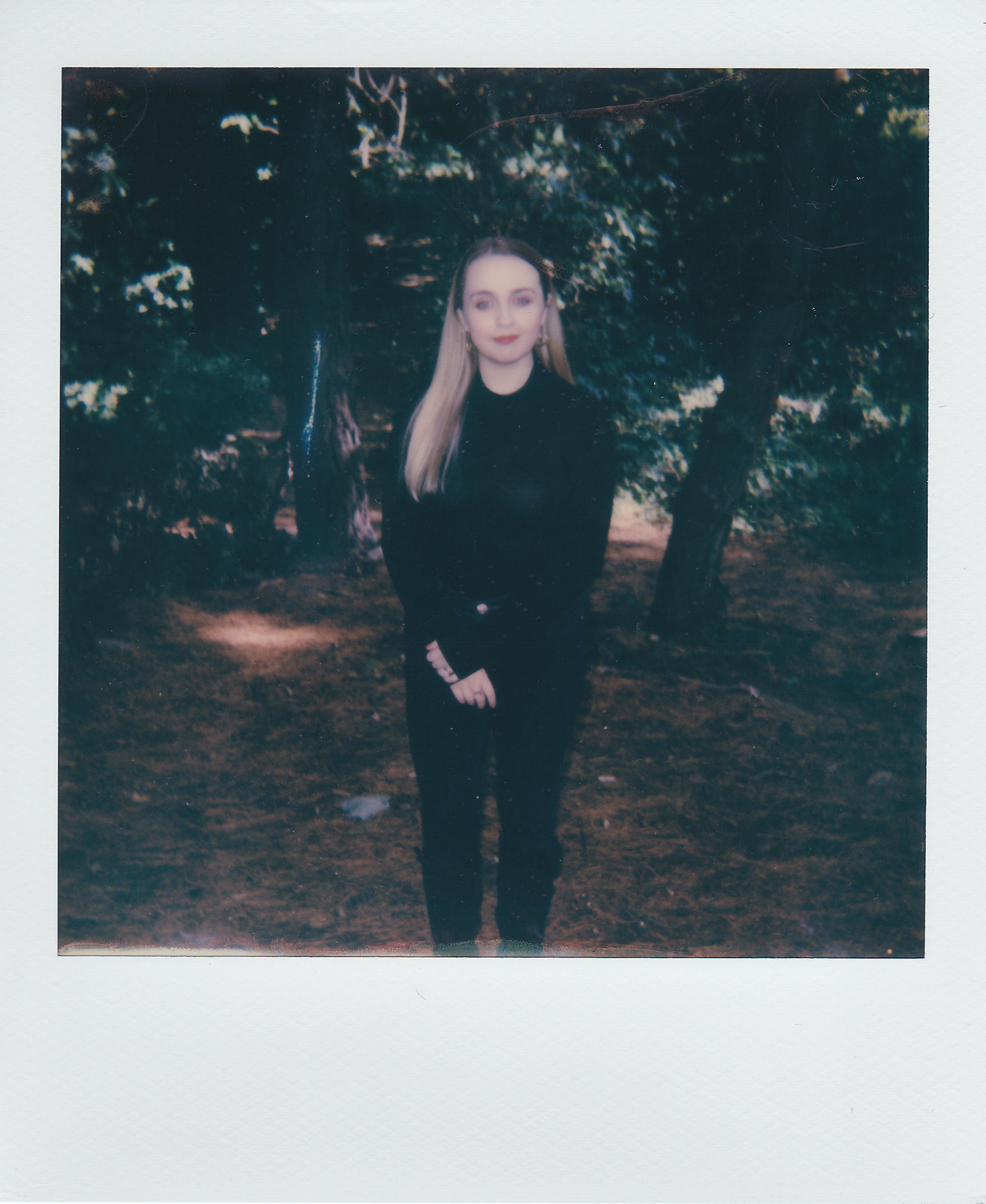 Blonde woman posing elegantly in a wooded area with a serene expression, creating a calm and natural atmosphere.