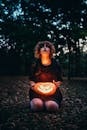 一名神秘女子跪在黑暗的公园里，举着一盏发光的南瓜灯，营造出怪异的万圣节氛围