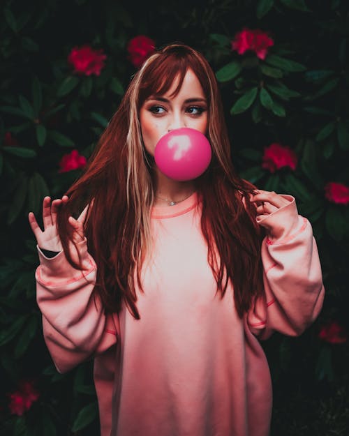 Dreamy woman in trendy clothes blowing pink gum in garden