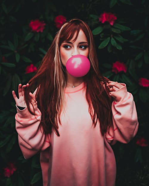 Dreamy woman in trendy clothes blowing pink gum in garden