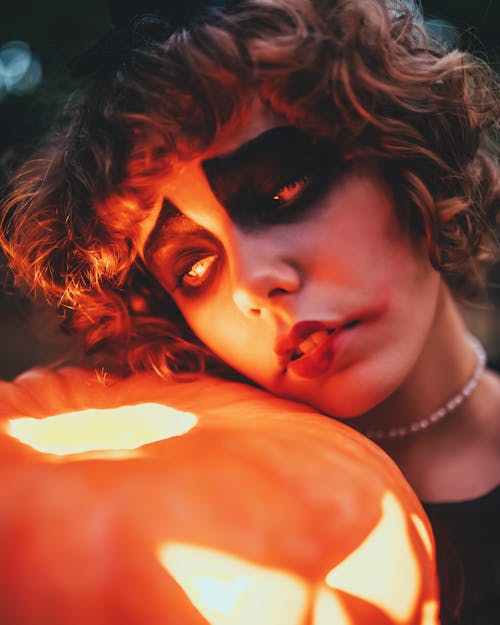 Crop sad female with terrible makeup and curly hair leaning on shiny carved pumpkin on Halloween night while looking away