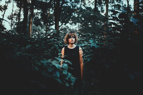 Young contemplative female with Halloween makeup in black wear looking up among trees in woods