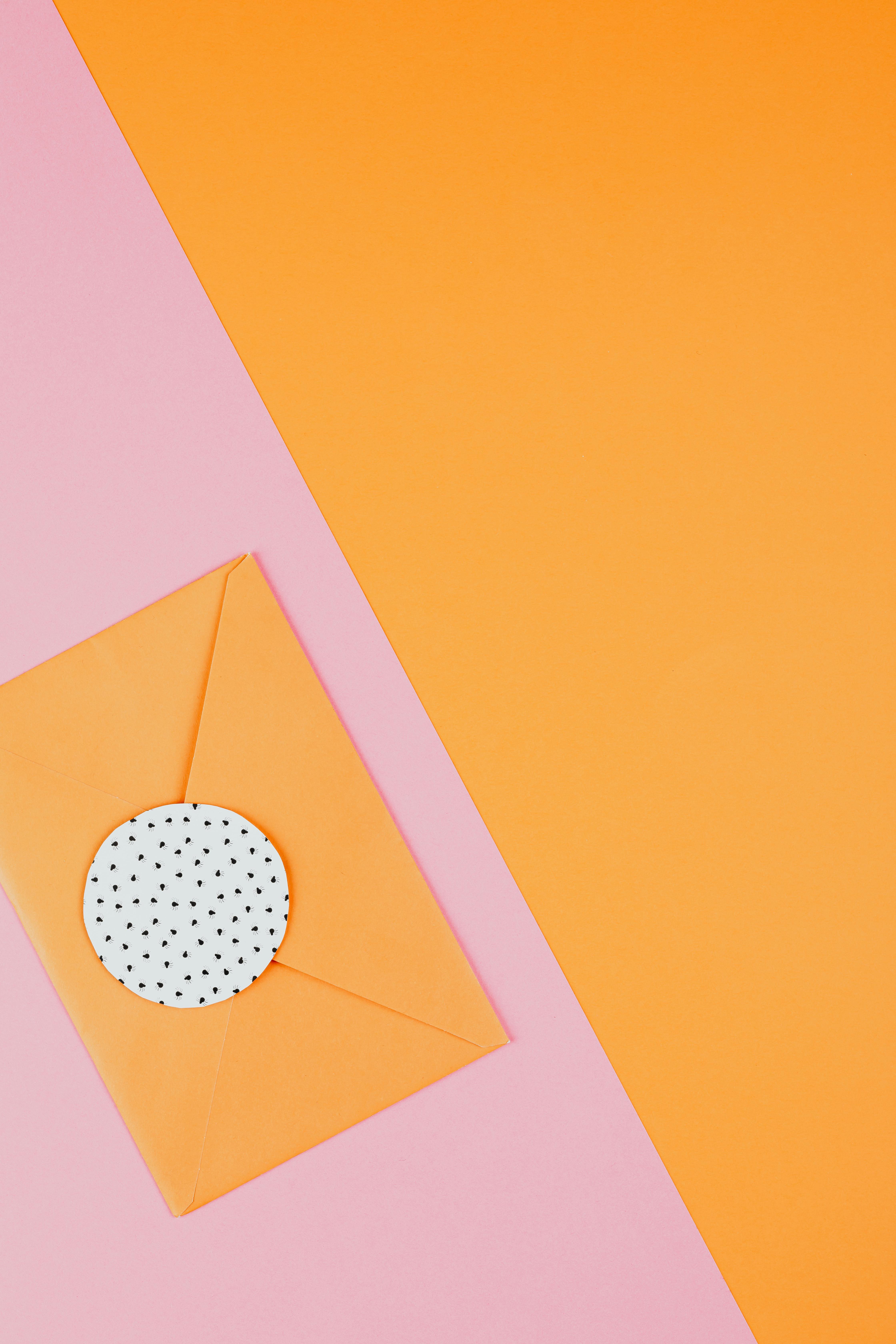 An unopened, carrot-orange-colored envelope is placed on overlapped sheets of orange and pink paper.
