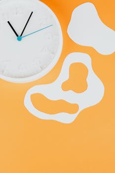 Flat lay of a white wall clock with abstract white paper cutouts on an orange background.