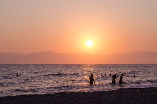 Free A beautiful sunset over a Greek beach with silhouettes of people swimming and playing in the sea. Stock Photo