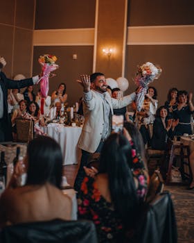 A joyful wedding scene with guests clapping and men holding flower bouquets indoors.