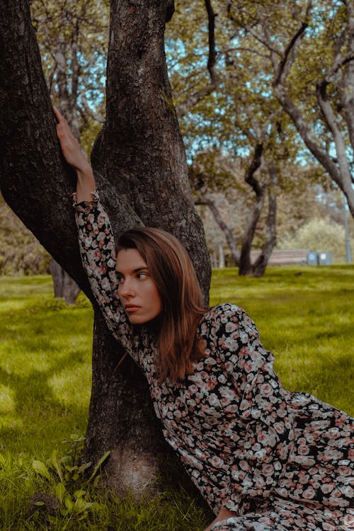 Free Young pondering female in stylish apparel leaning on tree while sitting on grass meadow in park and looking away Stock Photo