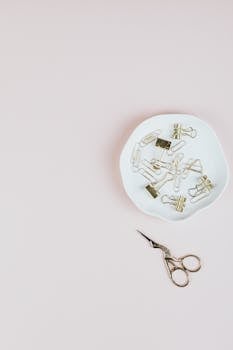 Top view of paperclips in dish and ornate scissors on a pale surface.
