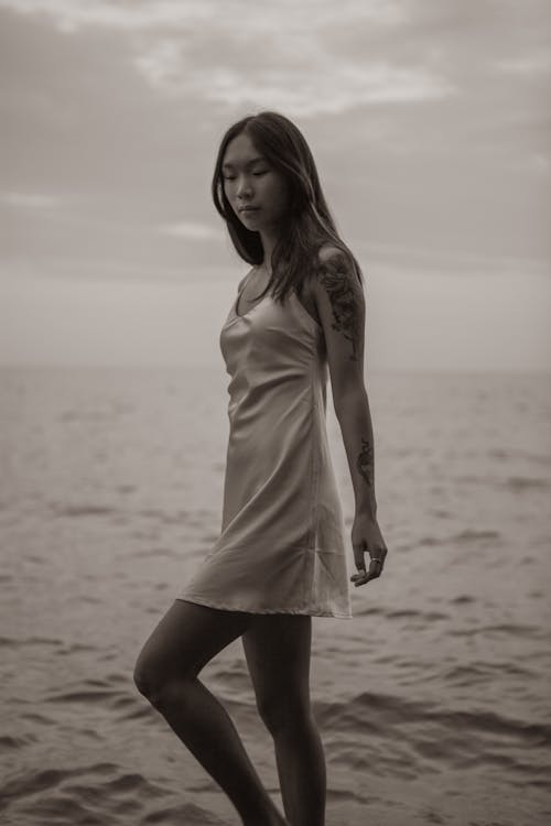 Free Side view of black and white young alluring Asian female in sundress walking along beach near waving ocean against cloudy sky Stock Photo