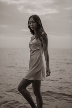 一位身穿太阳裙、年轻迷人的黑白混血亚裔女性，沿着海滩散步的侧面图，背景是波涛起伏的海洋和多云的天空