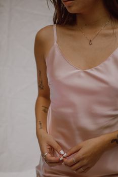 Crop anonymous young sensual woman with tattoos on arms in silk top standing against white background