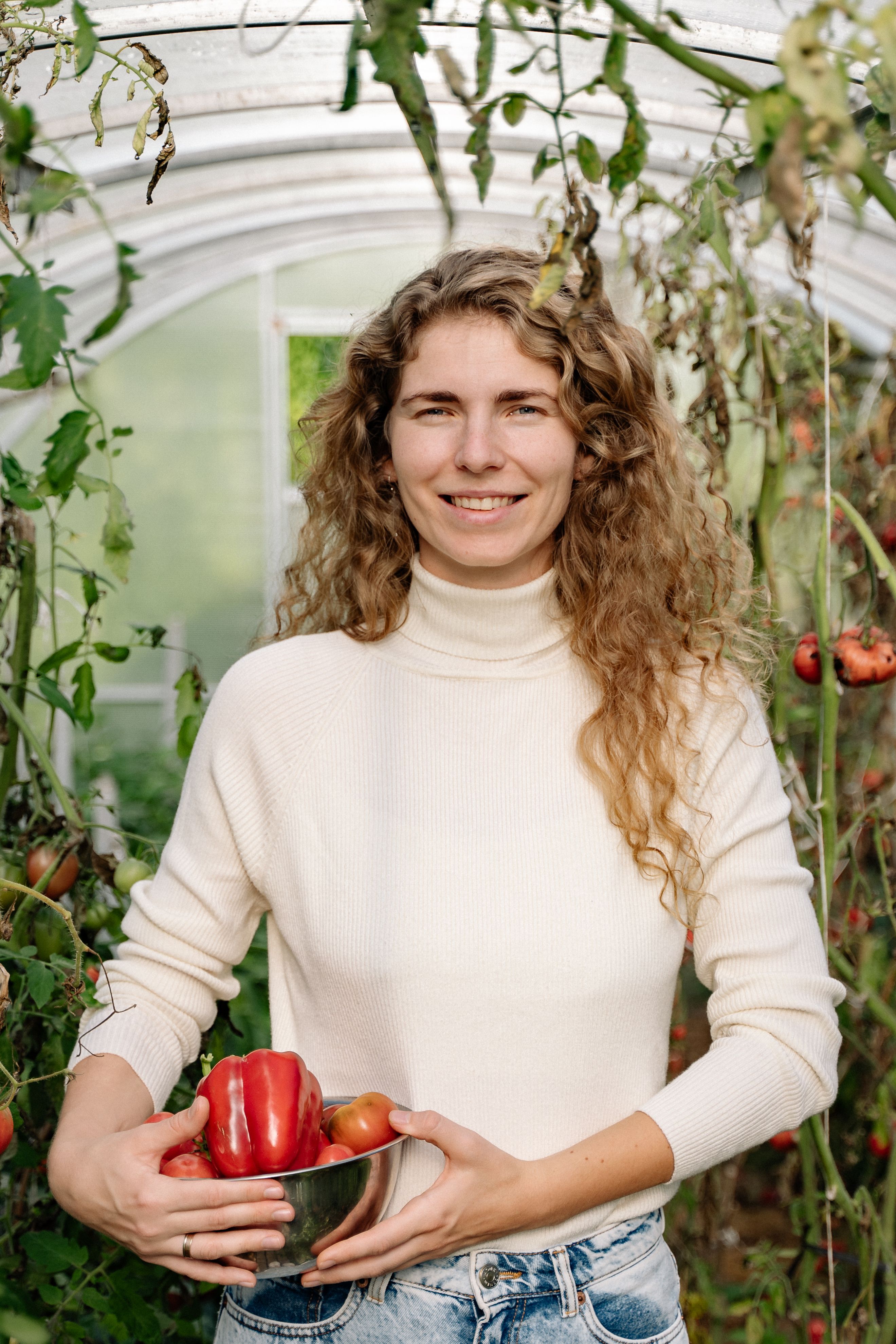 Caucasian woman with curly hair holding fresh bell peppers in a greenhouse.