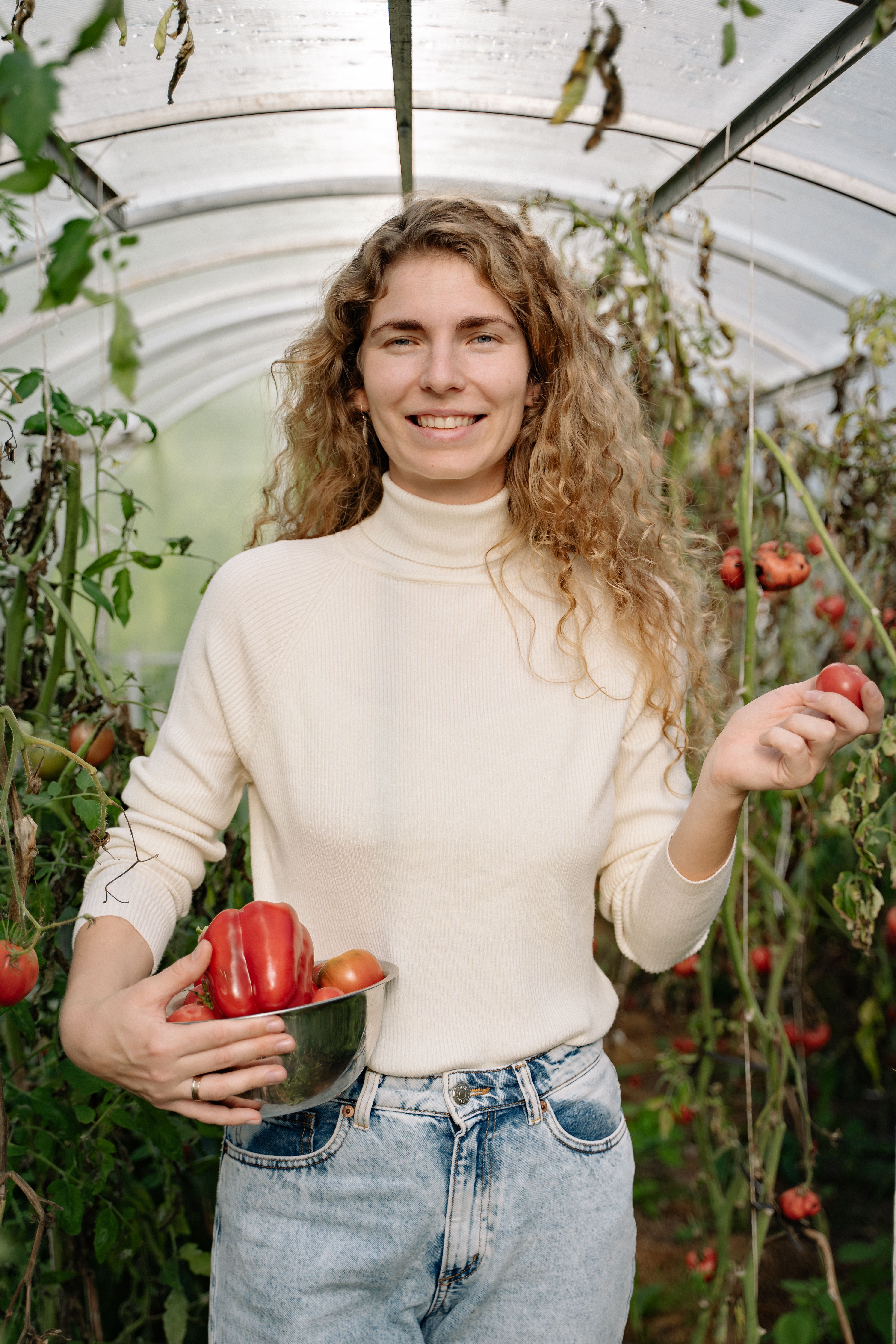 A woman holding a bowl of tomatoes and peppers in a lush greenhouse.