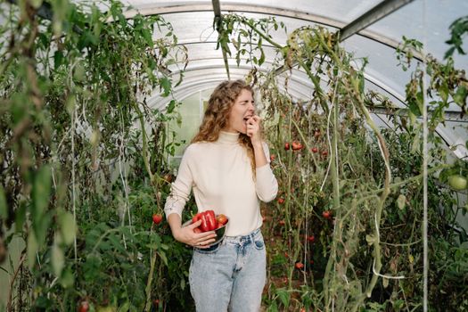 A woman enjoys freshly picked vegetables in a lush greenhouse setting.