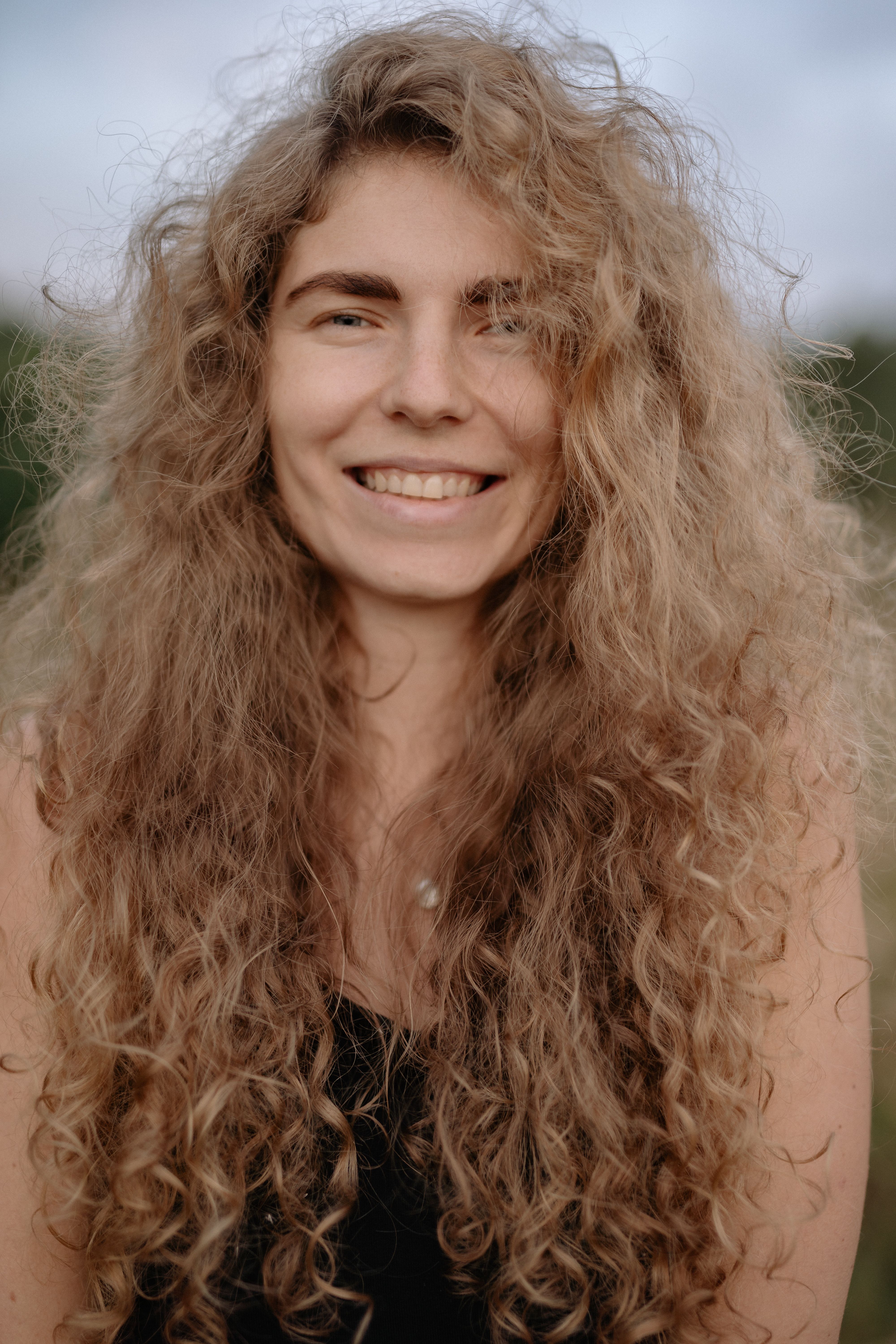 Close-up portrait of a smiling woman with long curly blonde hair against a natural background.
