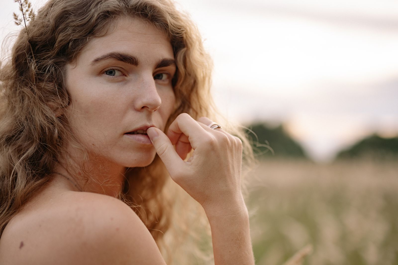 Woman with wavy hair outdoors in golden sunlight touching her lip thoughtfully.