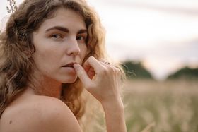Woman with wavy hair outdoors in golden sunlight touching her lip thoughtfully.