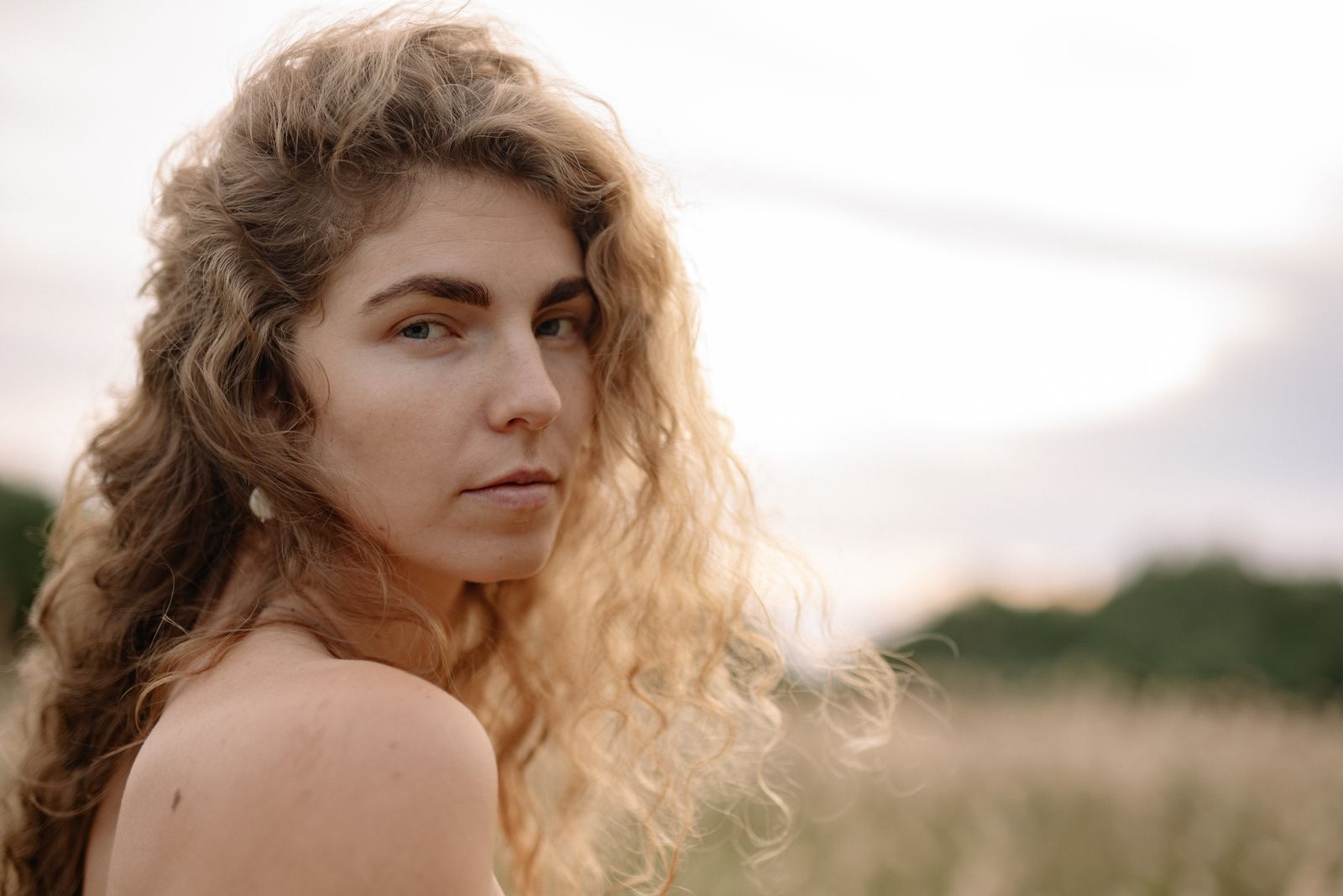 Woman with long wavy blonde hair looks over her bare shoulder in a field at golden hour