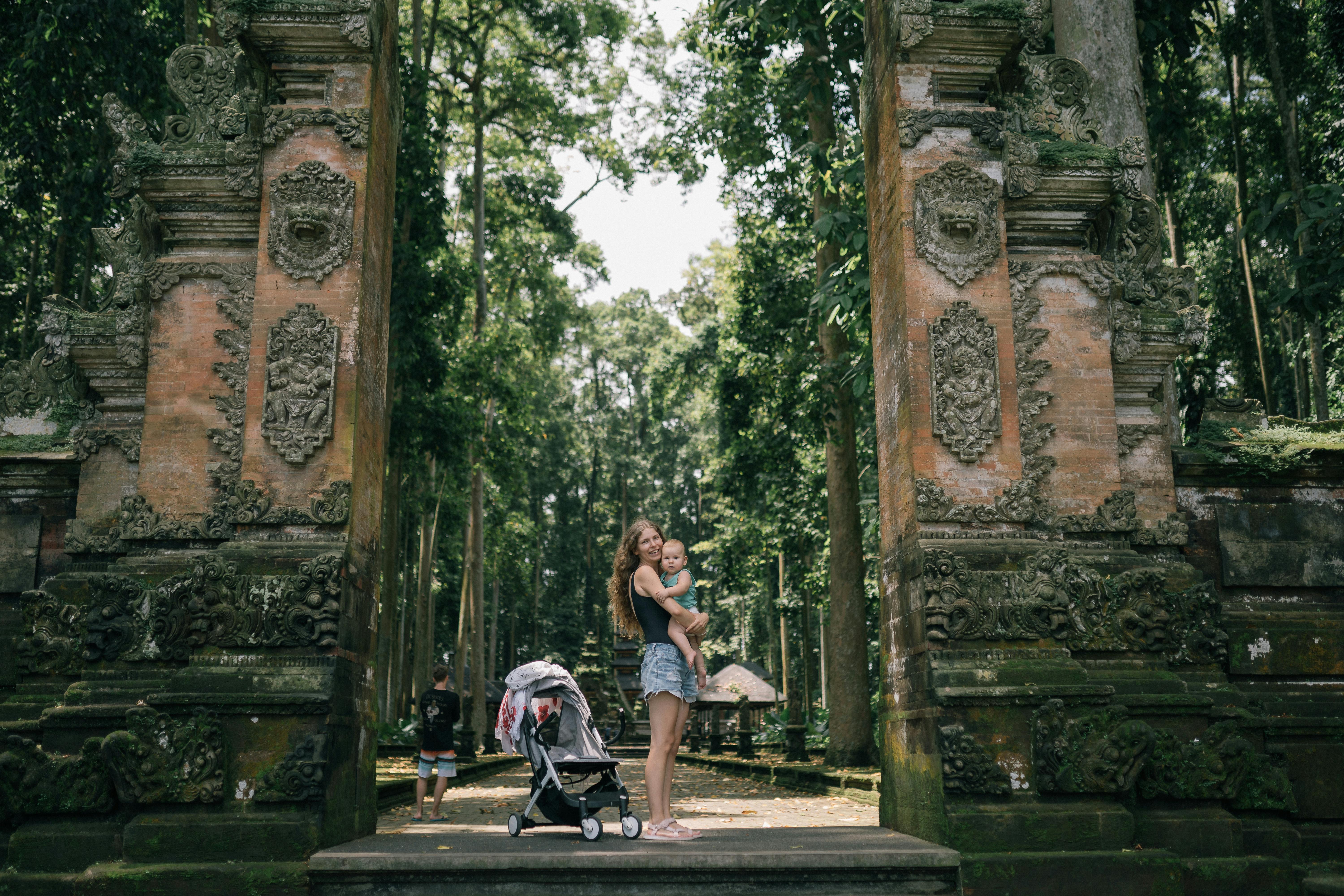 A mother holds her baby near an ancient temple gate surrounded by lush greenery.