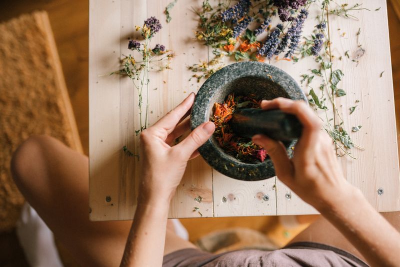 Hands using a mortar and pestle to grind colorful dried herbs on a wooden table.