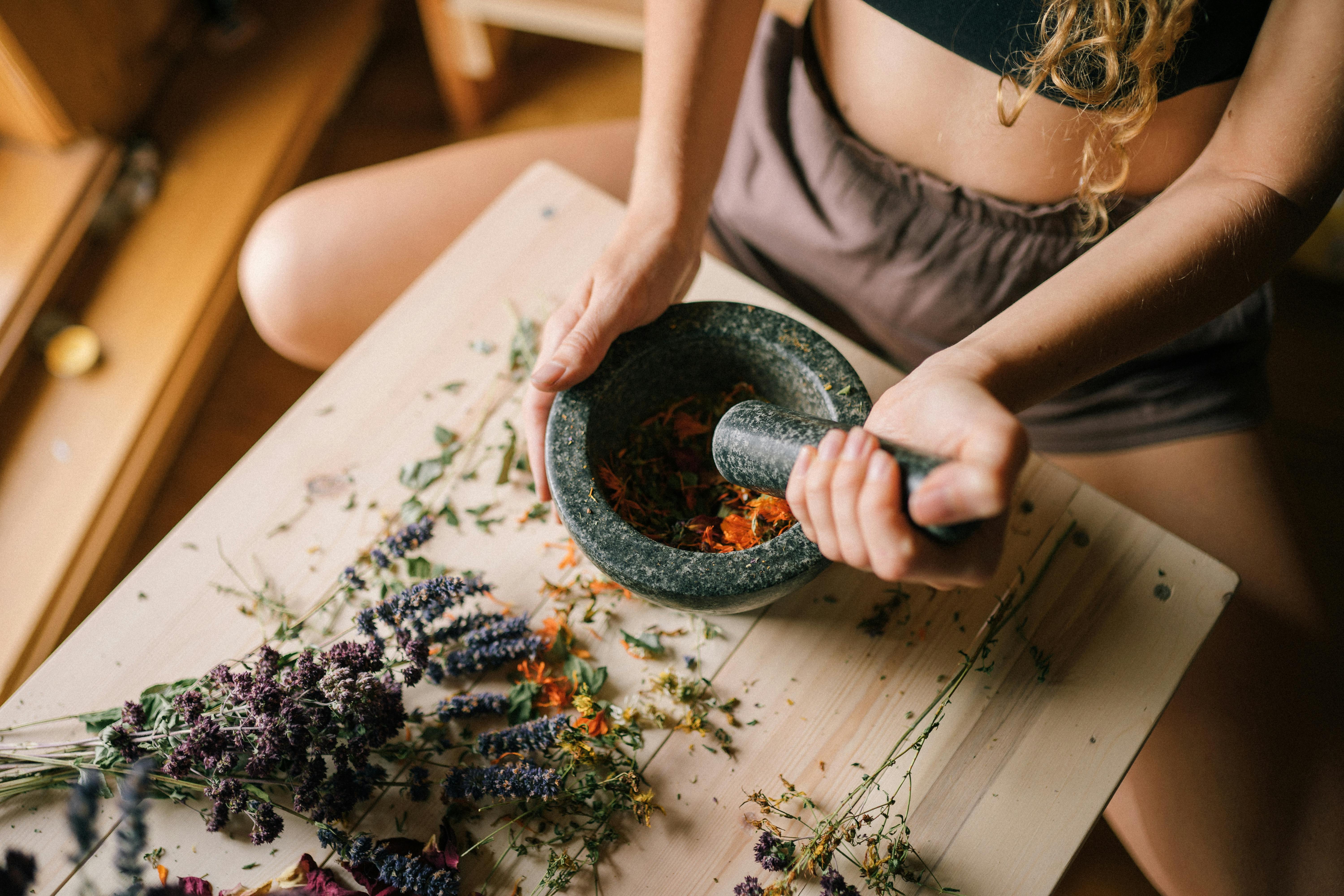 Close-up of hands using mortar and pestle to grind herbs, surrounded by dried flowers on wooden surface.